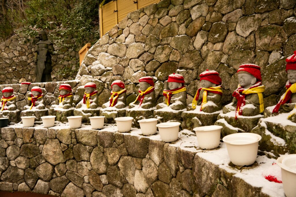 Fukuoka in Winter: Food, Illumination & Cozy Travel Row of Jizo statues with red hats and scarves on a snowy day in Fukuoka, Japan.
